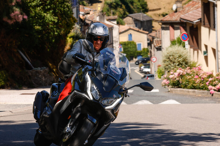 Un pilote sur un scooter Kymco CV3 noir circule sur une route de village en Occitanie avec les ruines d'un château médiéval en arrière-plan lors du Kymco Riders 2025.